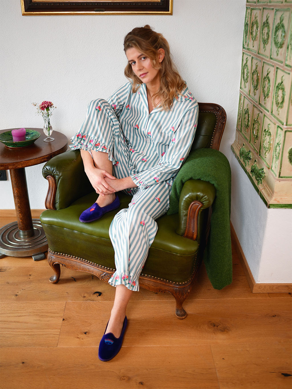 A woman sits in an olive green leather armchair wearing a luxury Nimo With Love light blue and white striped pajama set with pink floral prints and dark blue velvet slip-on shoes. The setting is a cozy room with wooden floors, a side table, and a green throw blanket draped over the chair.