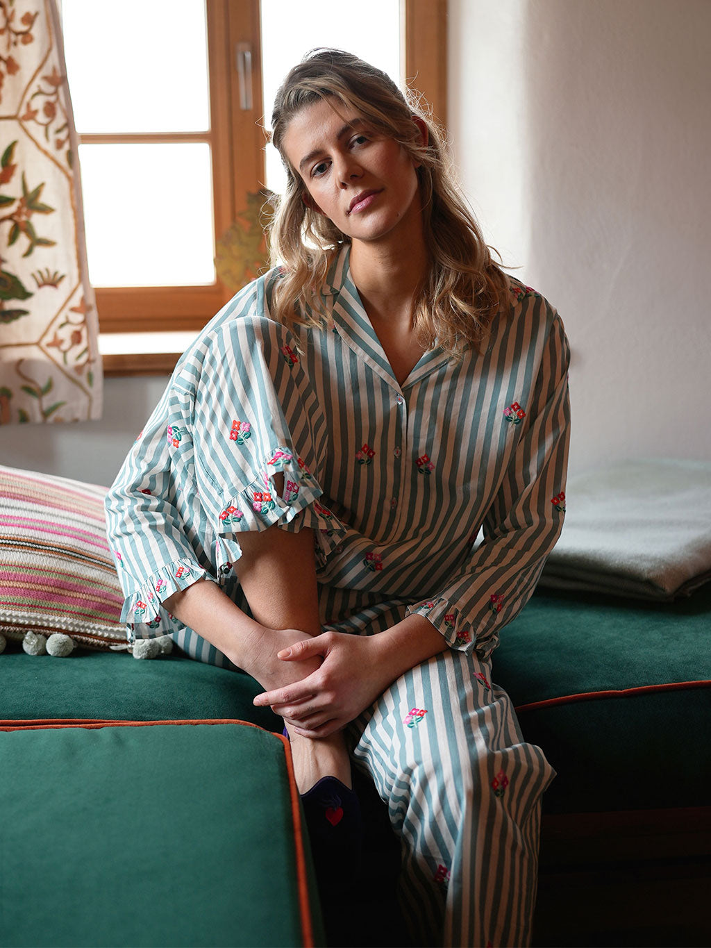 A woman wearing a luxury Nimo With Love matching striped pajama set with small embroidered floral details sits on a green sofa in a softly lit room.