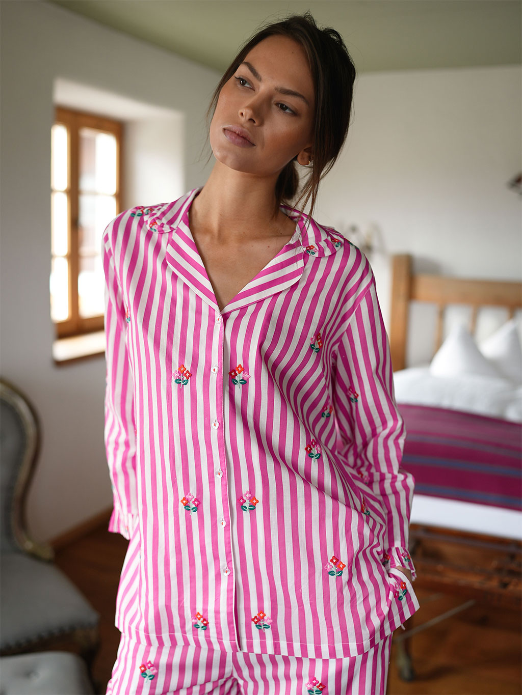 A woman wearing a luxury Nimo With Love pink and white vertically striped pajama set with small embroidered floral details, standing in a bedroom setting.