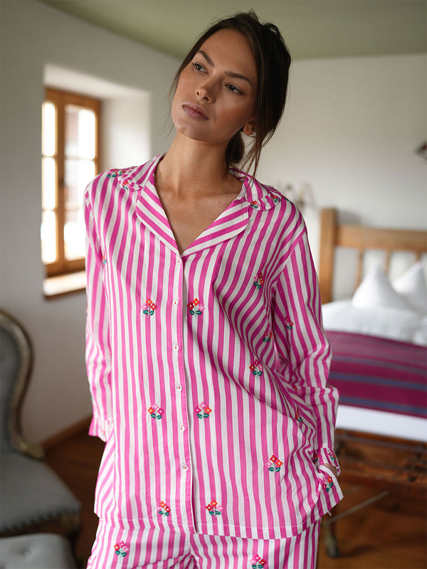 A woman wearing a luxury Nimo With Love pink and white vertically striped pajama set with small embroidered floral details, standing in a bedroom setting.