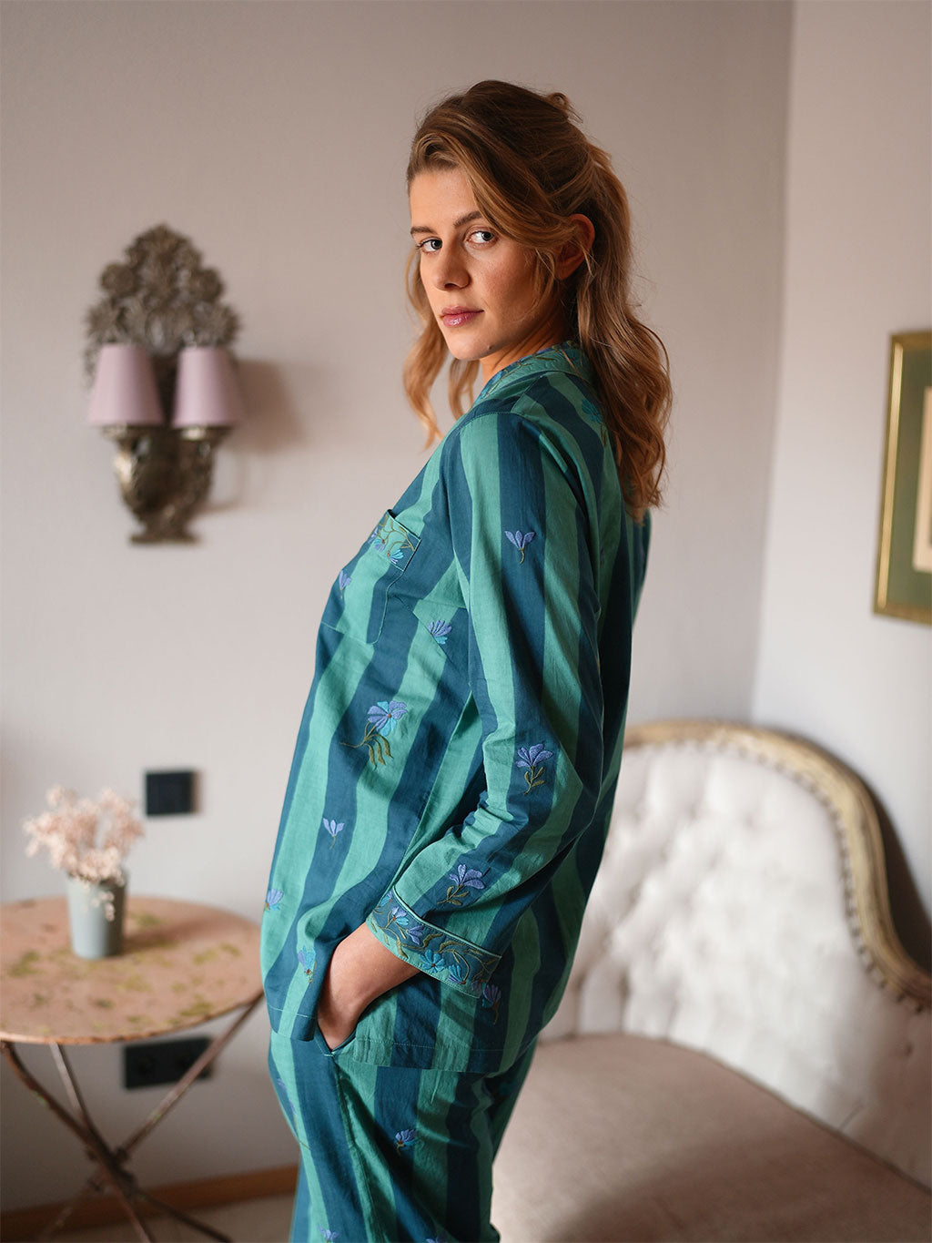 A woman wearing a luxury Nimo With Love teal and green striped pajama set with small embroidered floral details, standing in a softly lit bedroom.