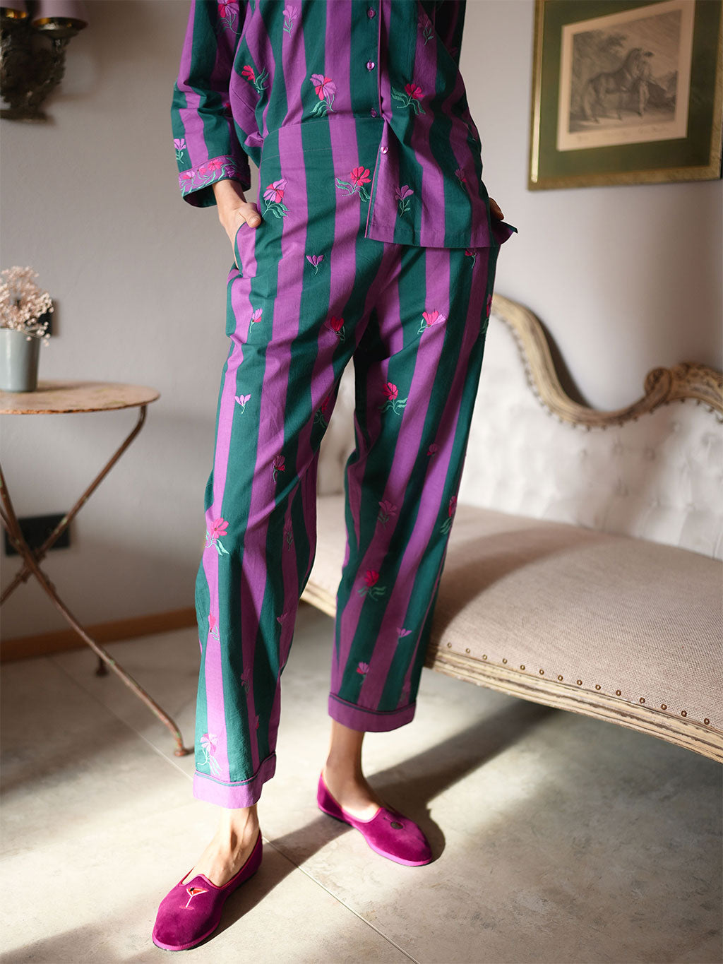 A model wearing a luxury Nimo With Love matching pajama set with vertical green and purple stripes and small pink floral motifs, paired with bright pink slip-on shoes, standing in a styled indoor room with a bench and framed wall art.