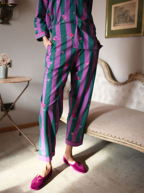 A model wearing a luxury Nimo With Love matching pajama set with vertical green and purple stripes and small pink floral motifs, paired with bright pink slip-on shoes, standing in a styled indoor room with a bench and framed wall art.