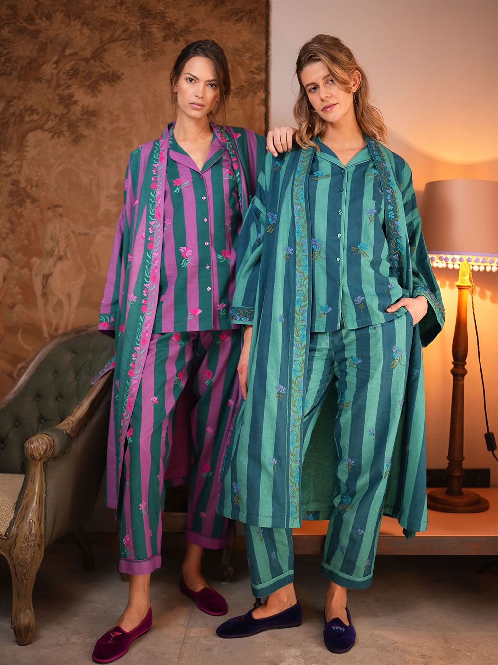 Two women modeling matching luxury Nimo With Love embroidered pajama sets in a warmly lit room. One set is purple and green with pink floral embroidery, and the other is teal and green with blue floral embroidery.