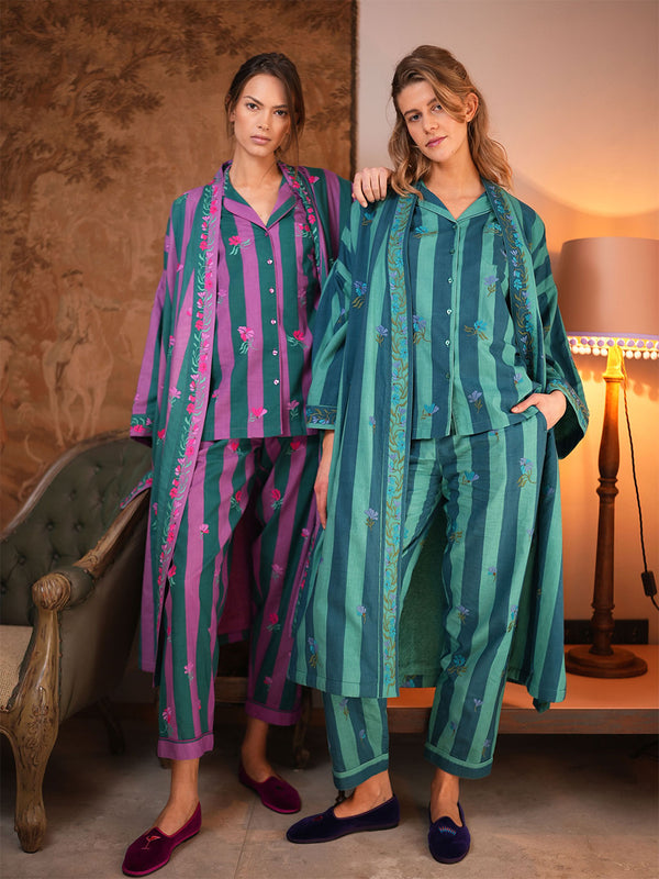 Two women modeling matching luxury Nimo With Love embroidered pajama sets in a warmly lit room. One set is purple and green with pink floral embroidery, and the other is teal and green with blue floral embroidery.
