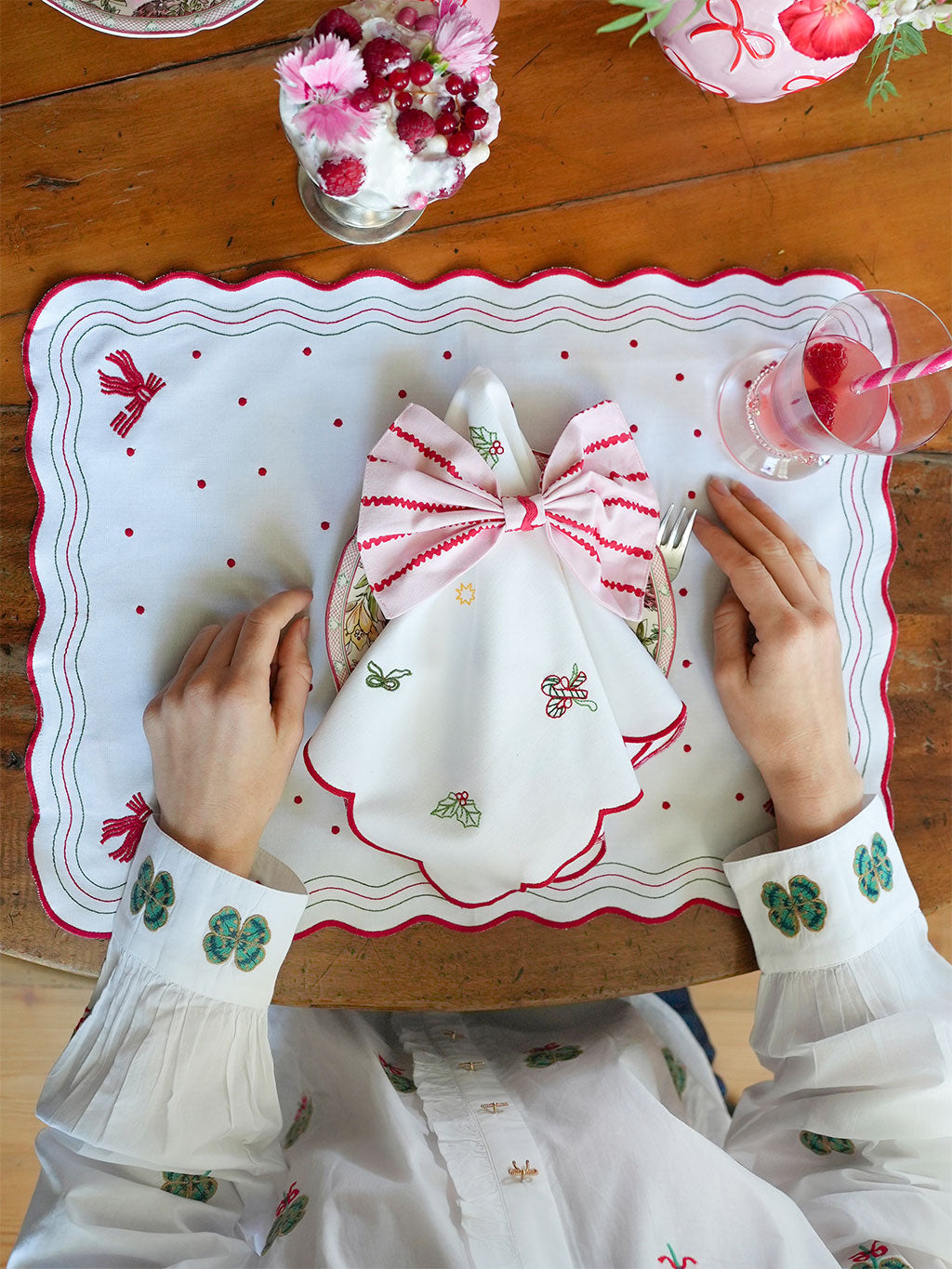 Person sitting at a Christmas table with a decorative placemat, holding a glass of red liquid.