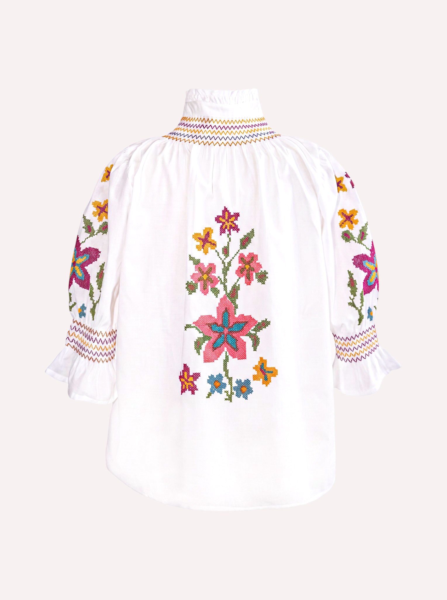 A white embroidered blouse with colorful floral patterns on the front and sleeves, featuring a high gathered neckline and ruffled cuffs, shown against a light beige background.