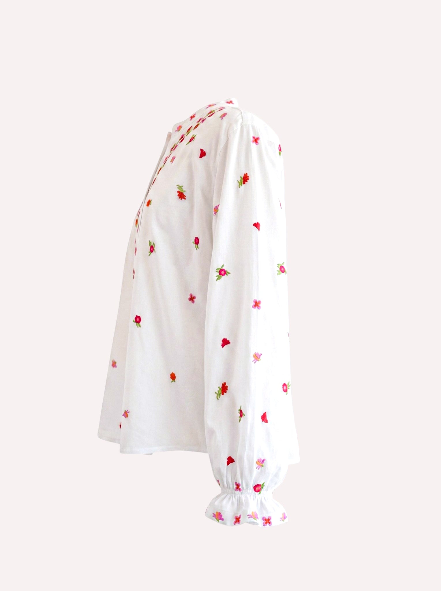 White Nimo With Love long-sleeve blouse with small red and green strawberry embroidery, featuring a gathered ruffled hem and cuffs, shown from a side profile on a light beige background.
