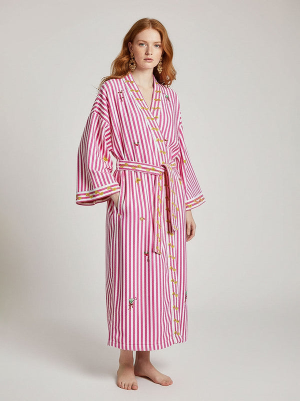 A woman modeling a luxury Nimo With Love pink and white vertically striped bathrobe with embroidered details, standing against a light gray studio background.