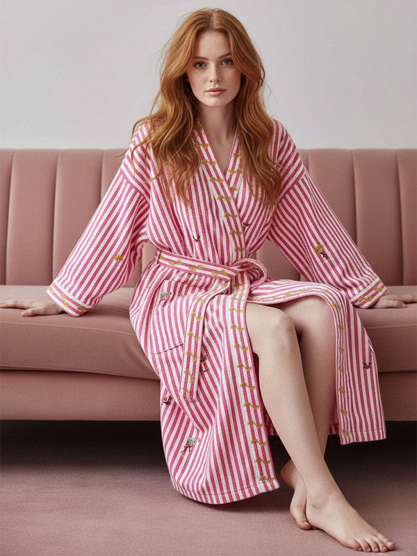 A woman with long red hair sits on a pink sofa wearing a luxury Nimo With Love pink and white striped bathrobe with embroidered details. The robe features a tie belt and decorative stitching.