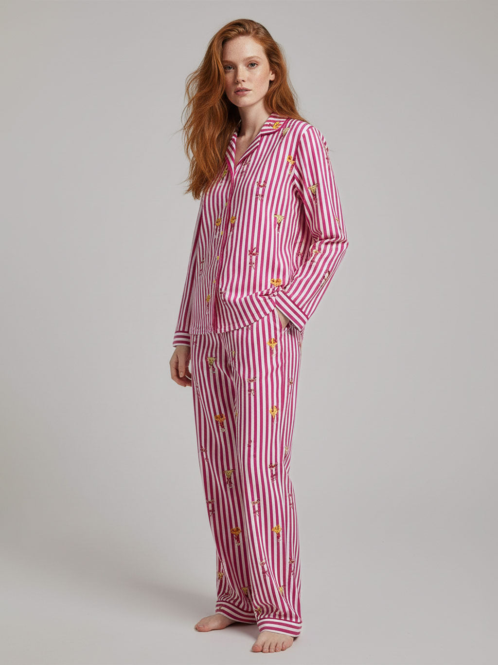 A woman modeling a matching two-piece pajama set with pink and white vertical stripes and small embroidered floral details, photographed against a plain light gray studio background.