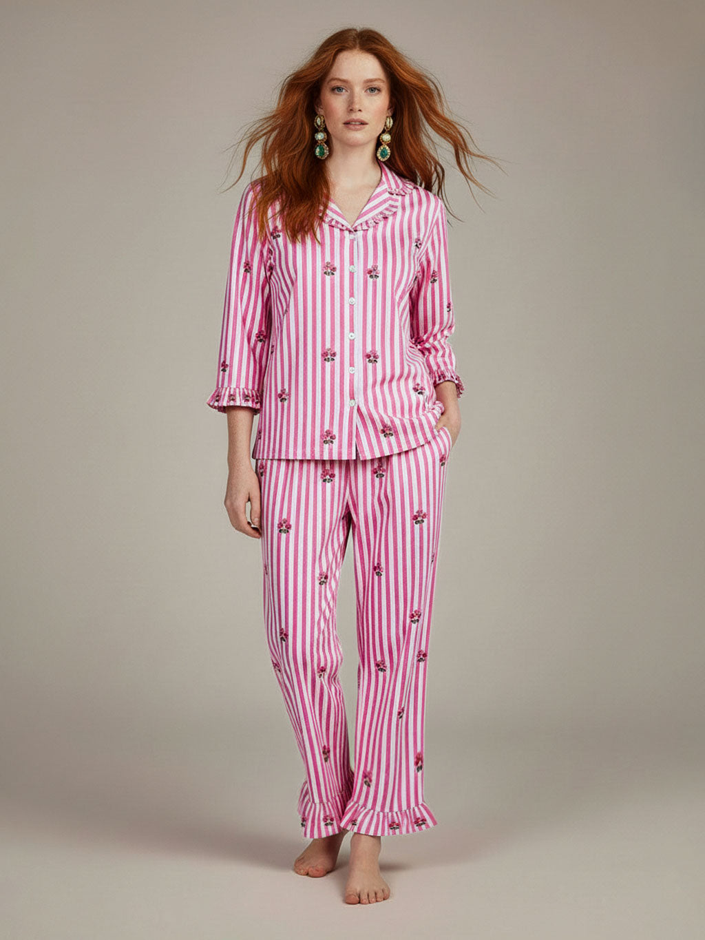 A woman modeling a luxury Nimo With Love pink and white striped pajama set with small cherry prints, standing against a neutral beige studio background.