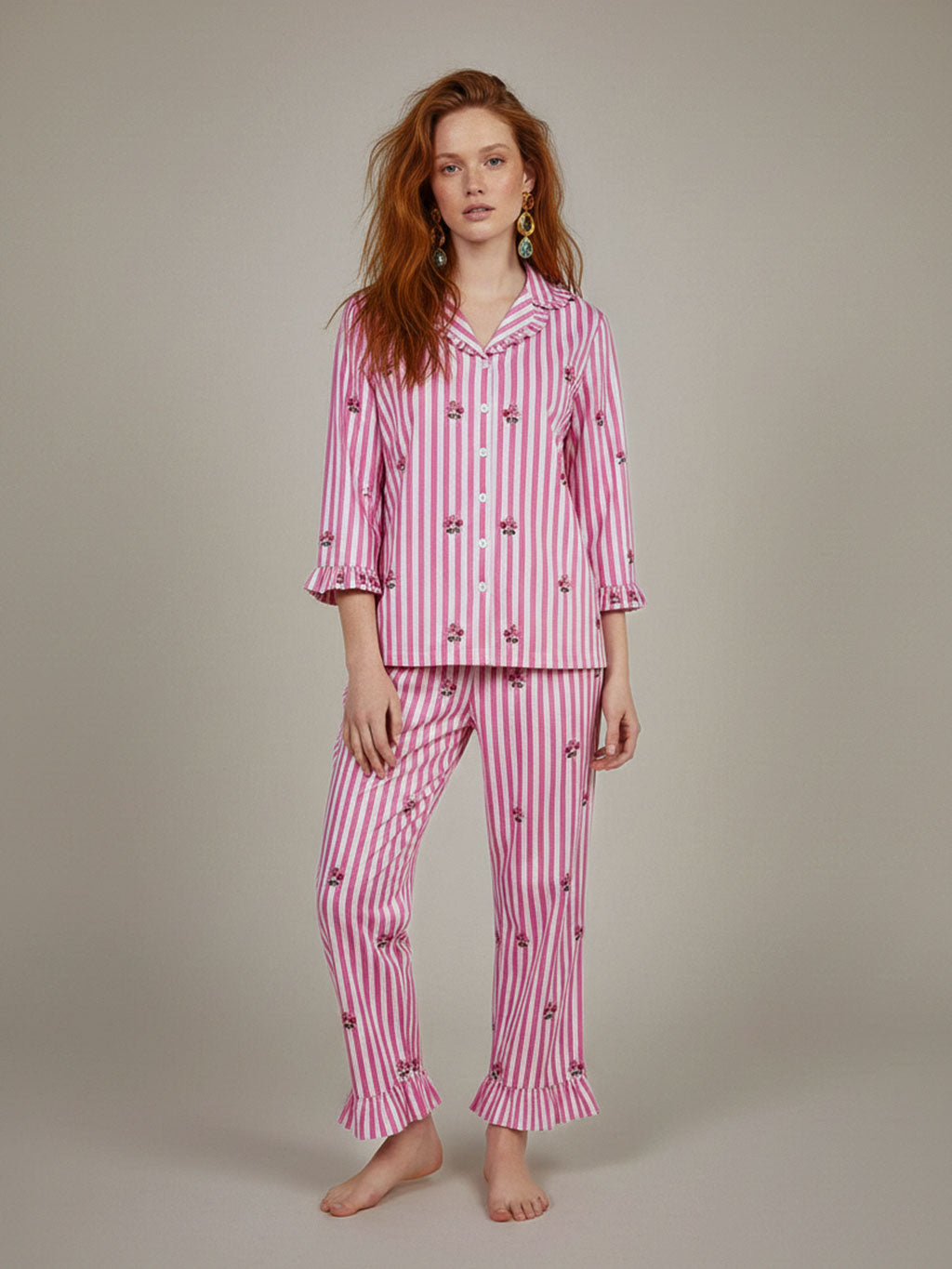 A woman modeling a luxury Nimo With Love two-piece pink and white striped pajama set with small embroidered floral details, standing against a neutral beige studio background.